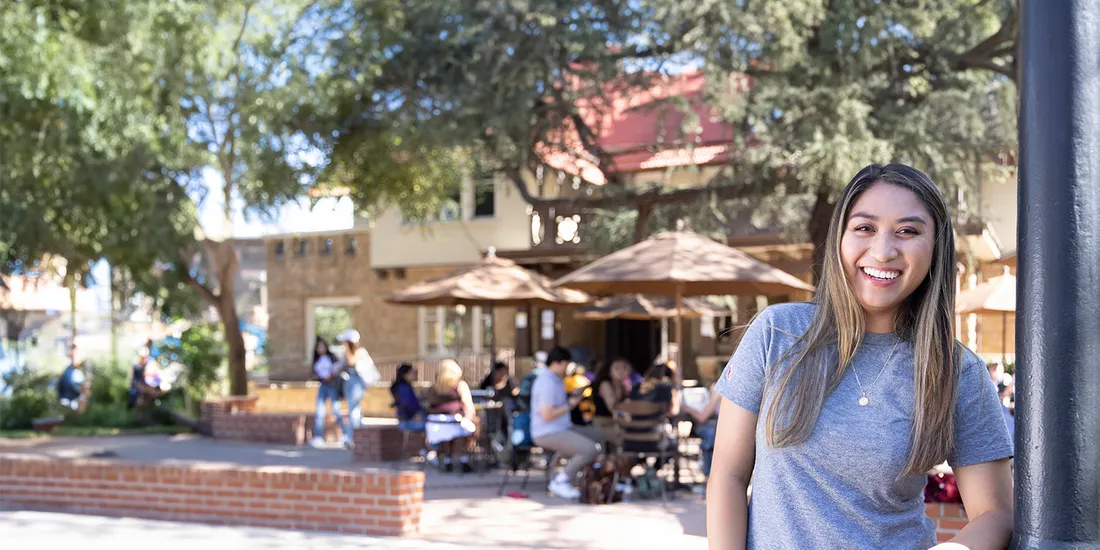 Female student smiling wearing an MSMU t-shirt, with students sitting and chatting on campus in the background.