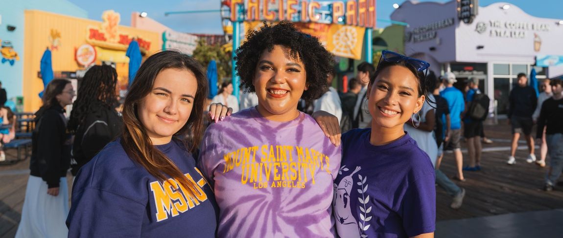 three students standing together in purple 91ɬ�� shirts smiling at the santa monica pier