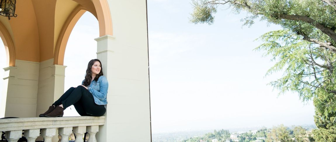 female student with brown hair sitting on a ledge in a jean jacket smiling