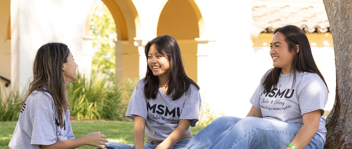 three female students in grey shirts smiling talking sitting on the grass