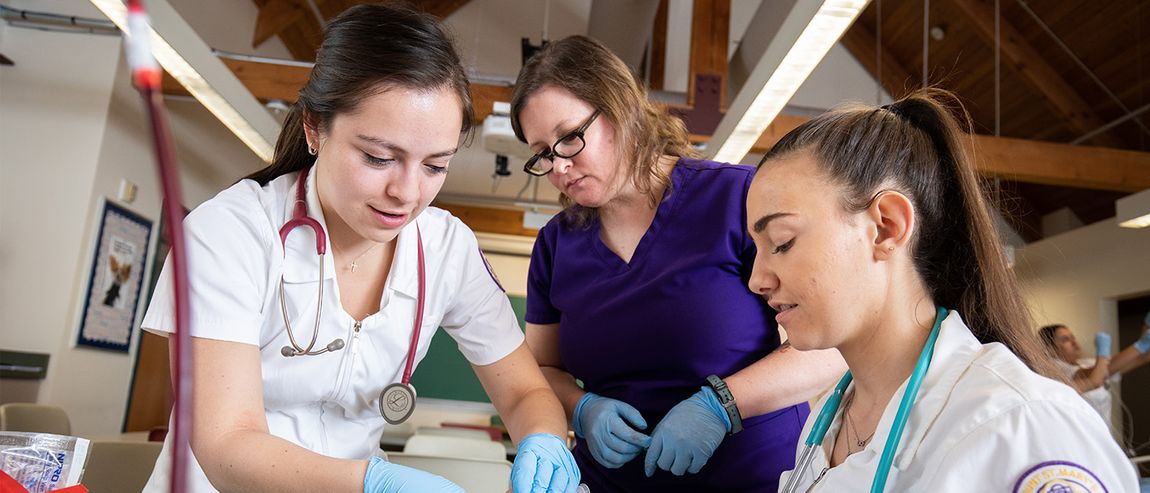 nursing students in white coats working on an IV while a professor watches
