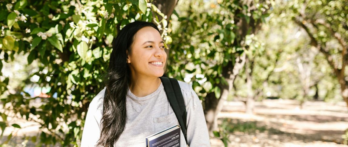 female student in white long sleeve with black backpack straps smiling looking right holding a book outside