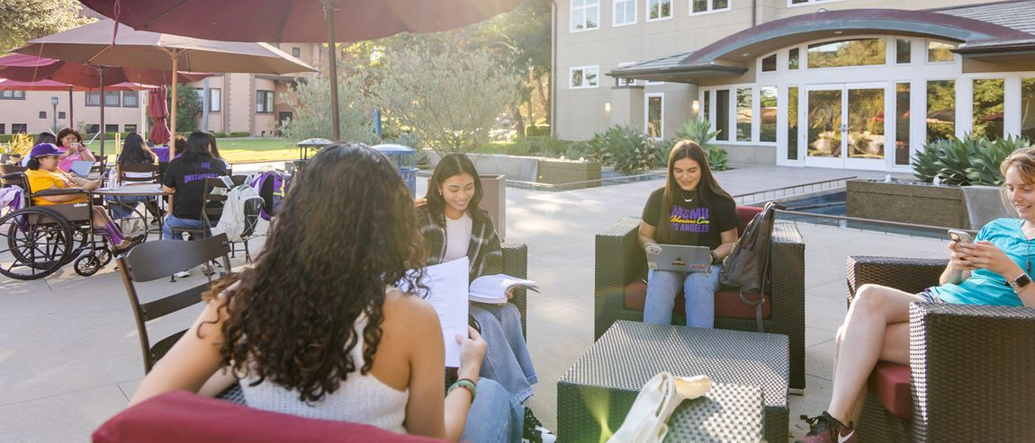 Students reading and studying while sitting under umbrellas in front of a multi-story building