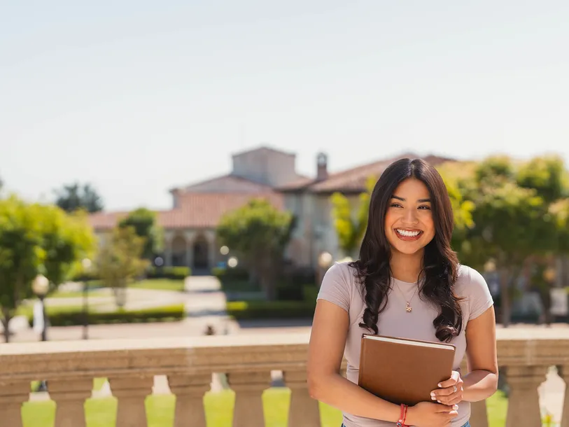 female student in light purple shirt smiling on chalon campus holding a book with trees and campus buildings in the background