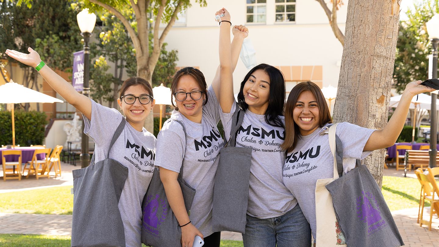 Four students cheering and smiling at the camera under some trees.