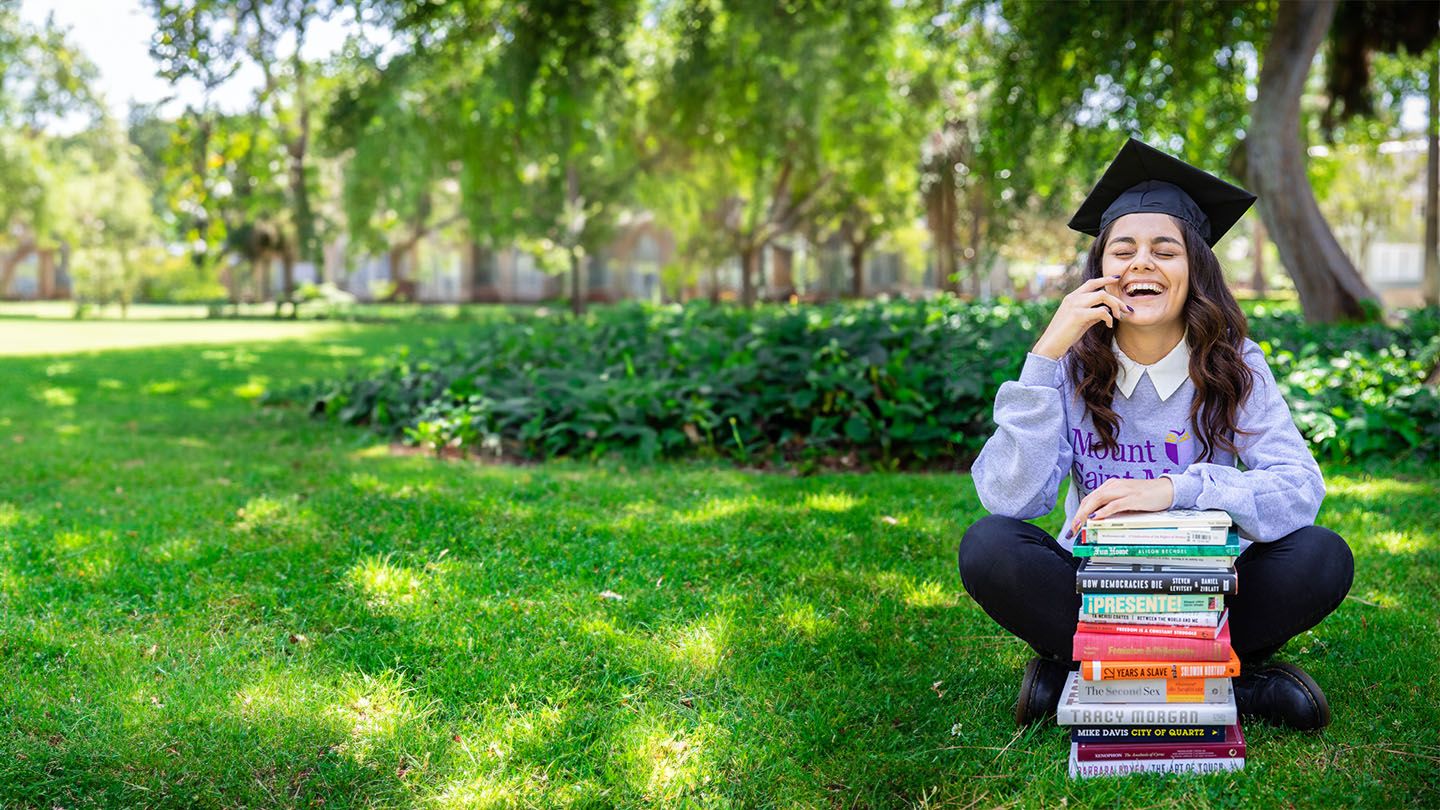 student sitting in grass laughing with a stack of political science books in front of her