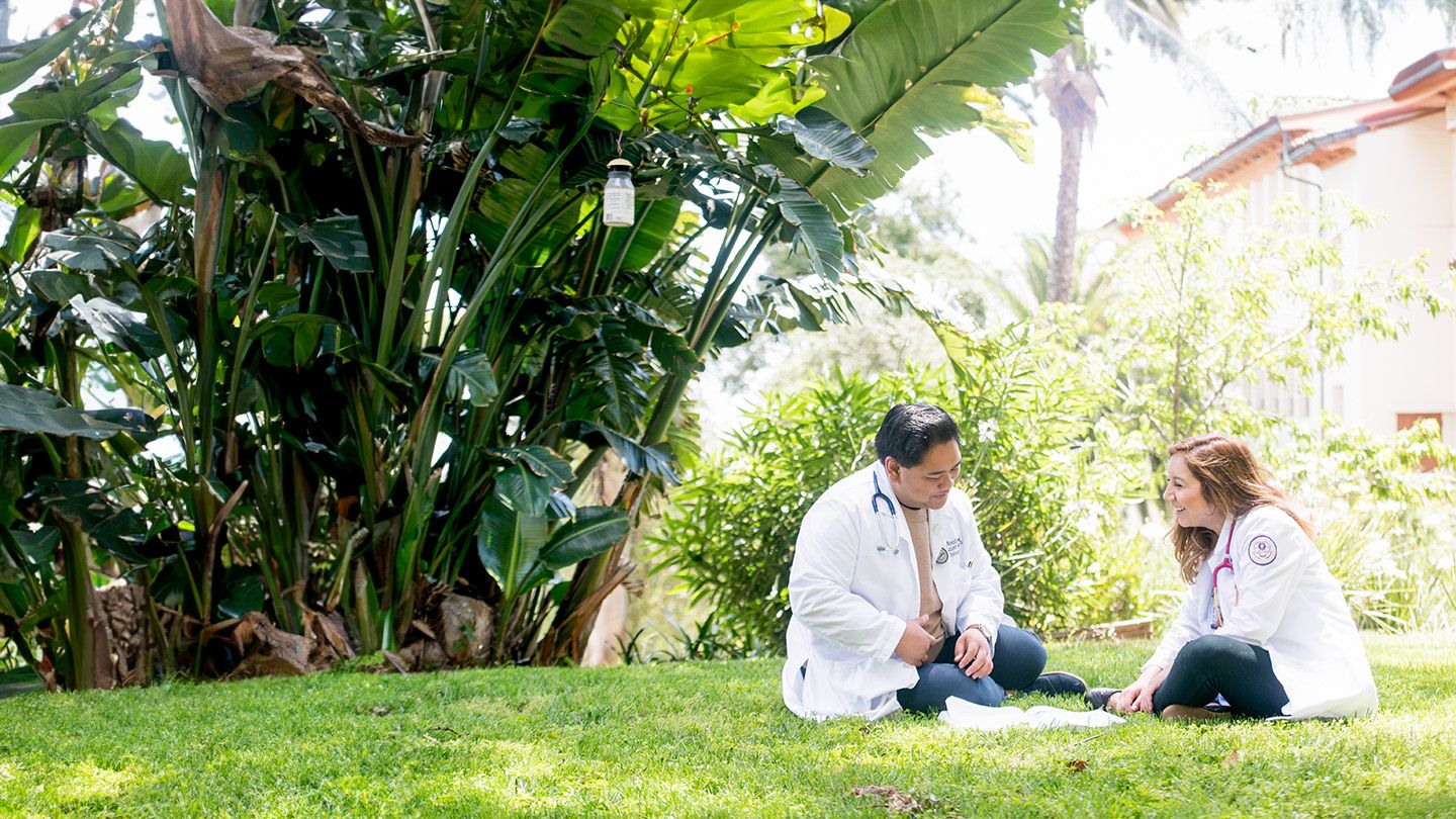 two nursing students in white coats sitting on the chalon campus grass talking