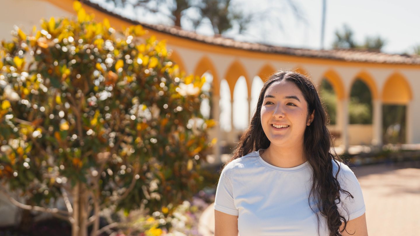 female student with long brown hair smiling walking outdoors with a corridor and some plants behind her