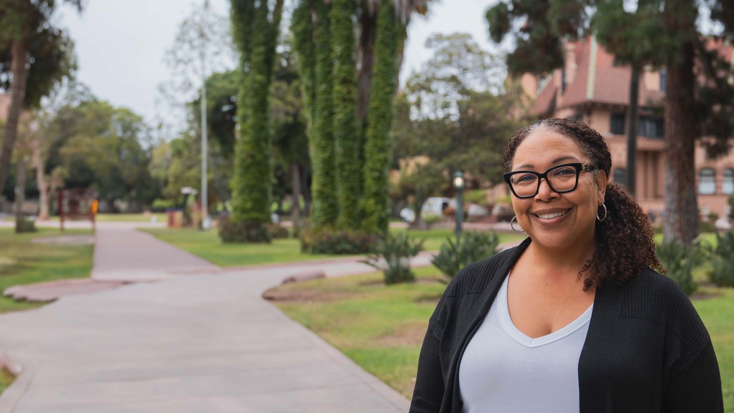 adult woman smiling, wearing black glasses and a black cardigan over a white shirt. She's standing outside, in front of a historical red-biege mansion and a green lawn.