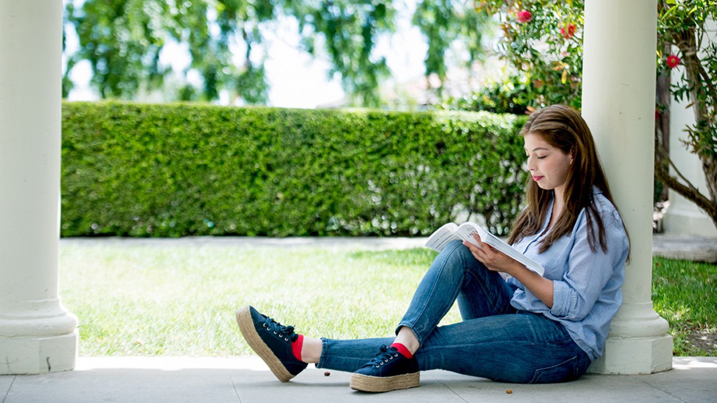 Young woman sitting and reading outdoors with a lawn and hedge in the background