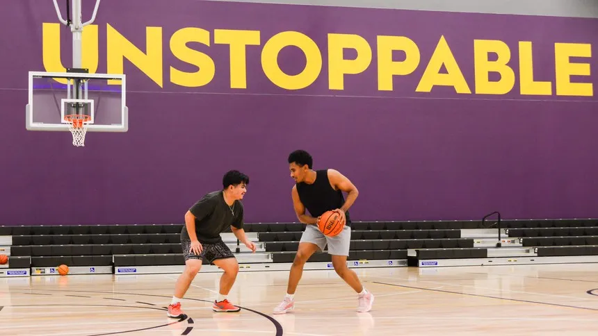 Two students play one-on-one basketball in the indoor gymnasium beneath a bold “UNSTOPPABLE” wall graphic.