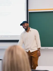 male student smiling talking in front of a projector screen in a classroom, wearing a baseball cap and white shirt with brown pants