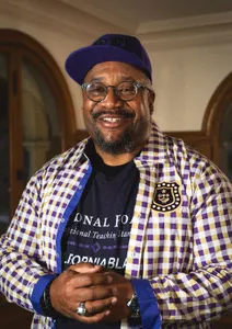 man in plaid shirt with black shirt underneath smiling, wearing glasses and a hat
