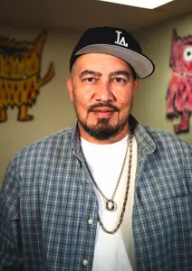 man in white shitr with blue long sleeve looking at camera, with a hat on