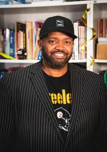 man in black suit jacket smiling with a hat on, behind him is a bookshelf filled with books
