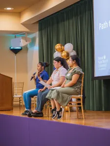 three female students sitting in chairs on stage, left one holding a mic as she speaks