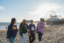 group of four female students walking and laughing on the beach, with the santa monica pier in the background
