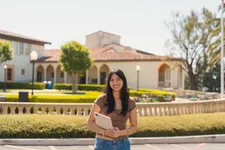 female student in brown shirt and jeans, holding an ipad to her chest smiling outdoors on the chalon campus