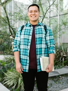 male student with teal plaid shirt on smiling standing outdoors with glass windows behind him