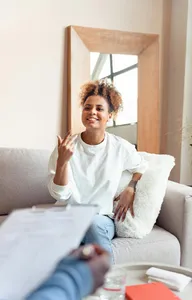 woman in white sweater and jeans sitting smiling on couch talking to a therapist who is holding a clipboard