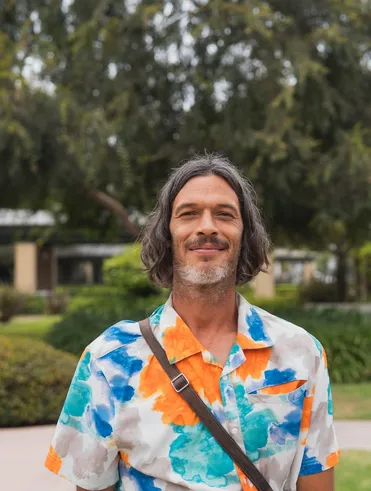 man with longer hair in a colorful blue and orange shirt with a strap around his should smiling outdoors