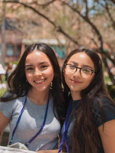 two female students with long dark hair smiling standing together, left one wearing a grey shirt the right wearing a black shirt