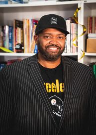 man in black suit jacket smiling with a hat on, behind him is a bookshelf filled with books