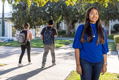 female student in cobalt blue shirt and long brown hair smiling, with two students with backpacks walking away in the background