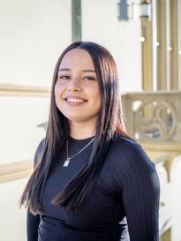 female student in black long sleeve and long brown hair smiling in front of beige building