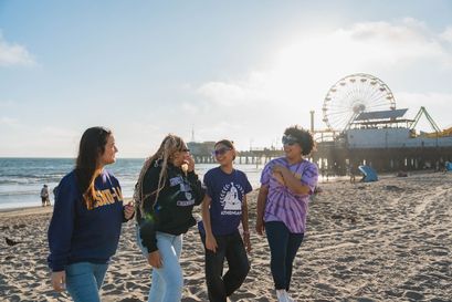 group of four female students walking and laughing on the beach, with the santa monica pier in the background
