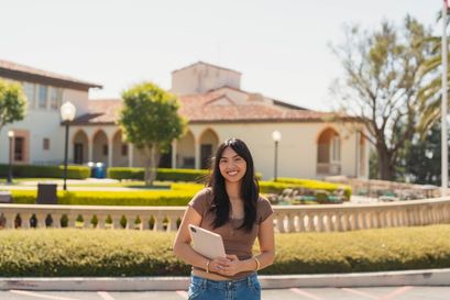 female student in brown shirt and jeans, holding an ipad to her chest smiling outdoors on the chalon campus