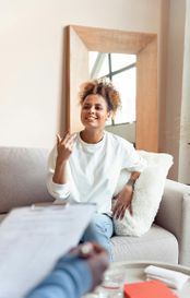 woman in white sweater and jeans sitting smiling on couch talking to a therapist who is holding a clipboard
