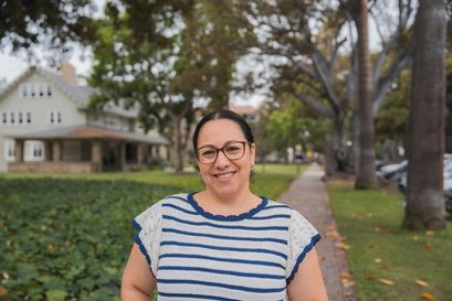woman in white and blue stripped blouse and glasses smiling outdoors with some greenery and trees behind her