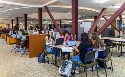 Students sitting at tables using computers and reading in a large room in a library.