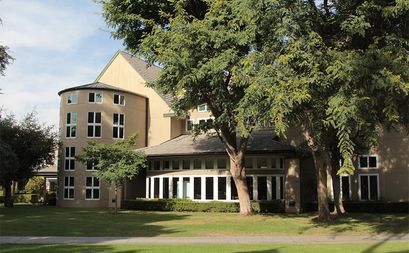 A rounded building with large windows seen through tall trees.