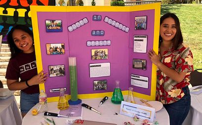 Two students smiling on either side of a posterboard display which contains information about the WISH Club at ˮ����