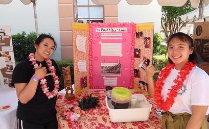 Two students wearing leis and smiling in front of a poster board with information about the Polynesian Culture Club on it.
