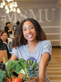 A student holding a basket of vegetables and smiling at the camera at an ÿ�մ������ farmers market