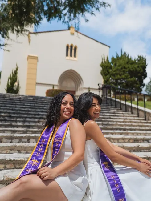 two female students sitting back to back on outdoor steps in front of the campus chapel smiling in white dresses and purple graduation stoles