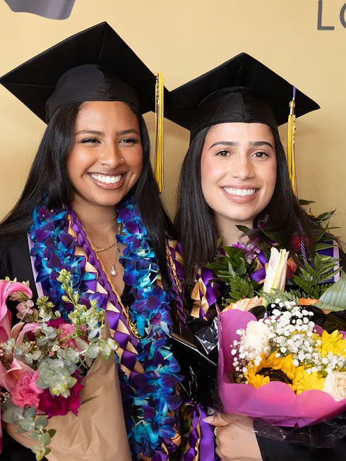 Two graduates smiling at commencement holding flowers