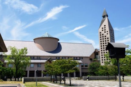 A short cream-colored building next to a tall tower with a tree-lined walkway leading to them.