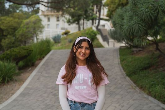 female student with brown long hair and white headband smiling outdoors in pink msmu shirt with white long sleeve shirt underneath and jeans