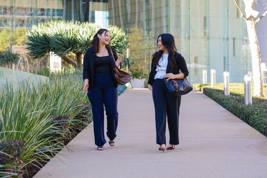 two women talking and walking outdoors in business attire in front of a glass building