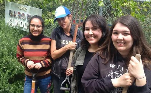 Some students holding gardening tools and smiling at the camera in front of a vine-covered fence