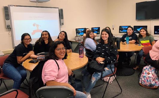 A group of students sitting around tables in a classroom and smiling at the camera