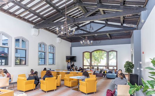 Students sitting in large gold-colored armchairs in a large study hall with tall windows at one end.