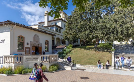 Students walking in front of a large building and a grassy hill covered with trees