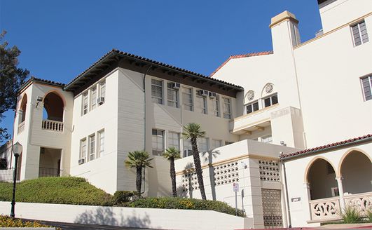 A large multi-level white building with a small driveway wrapping around lined with hedges.