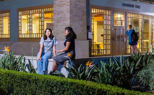 Two students sitting on a low wall and talking in front of a large building