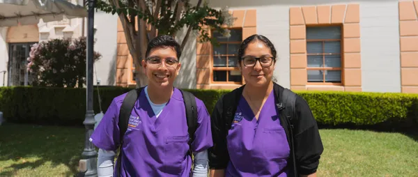 two nursing students smiling while standing outdoors and wearing scrubs